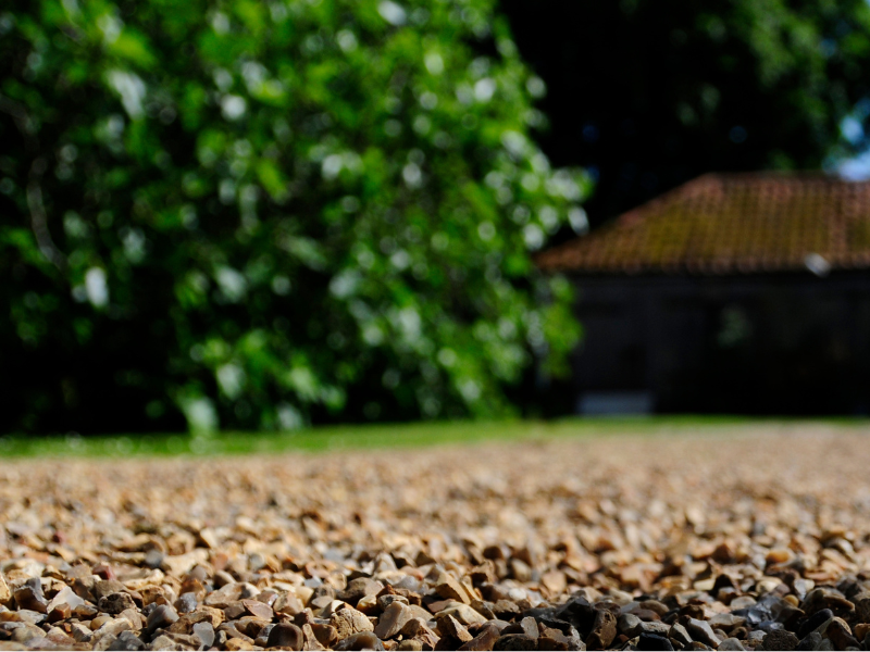 Close-up of gravel driveway with blurred green trees and a brick outbuilding in the background.